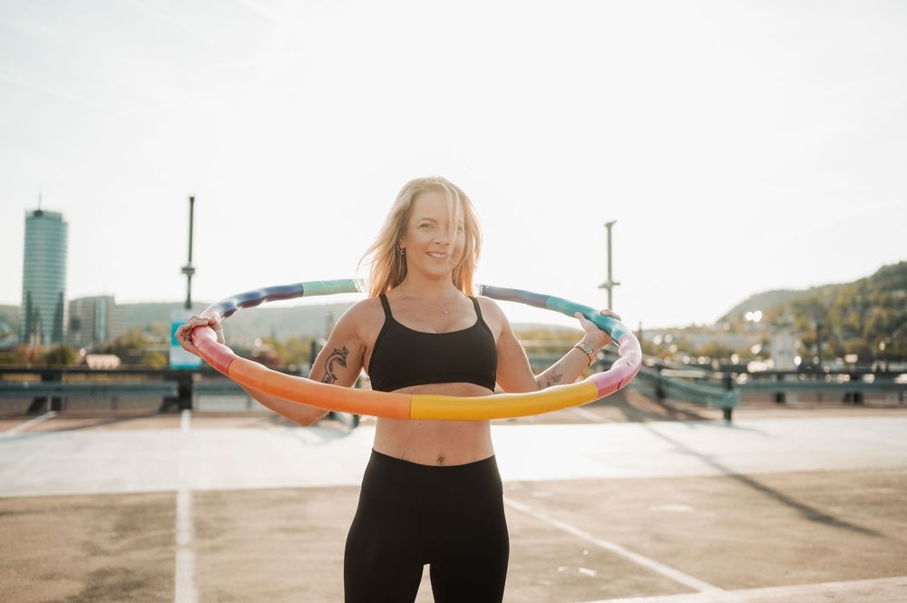 Person in athletic wear exercising with a hula hoop on an outdoor rooftop with city views in background.