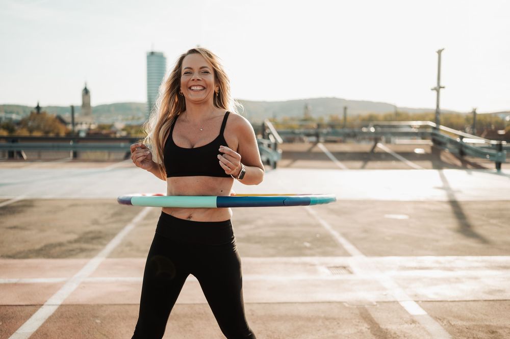 A person in black athletic wear demonstrates hula hoop exercises on a rooftop at sunset.
