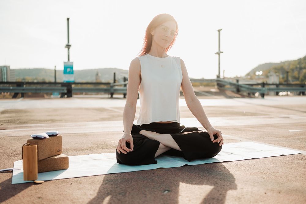 Person in workout attire meditating on yoga mat outdoors with water bottles nearby.