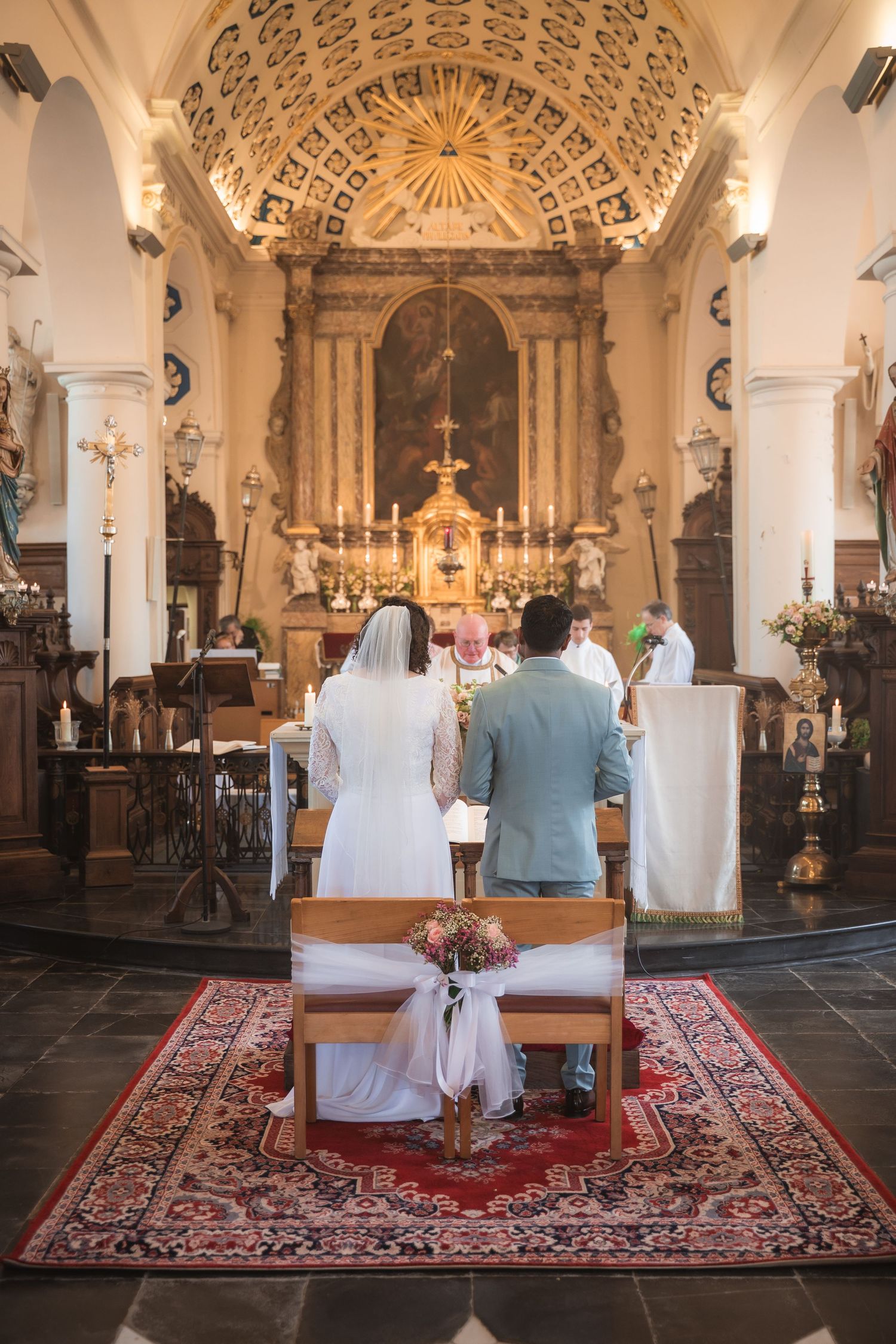 Bride and groom kneeling at church altar during traditional Catholic wedding ceremony with ornate decor.