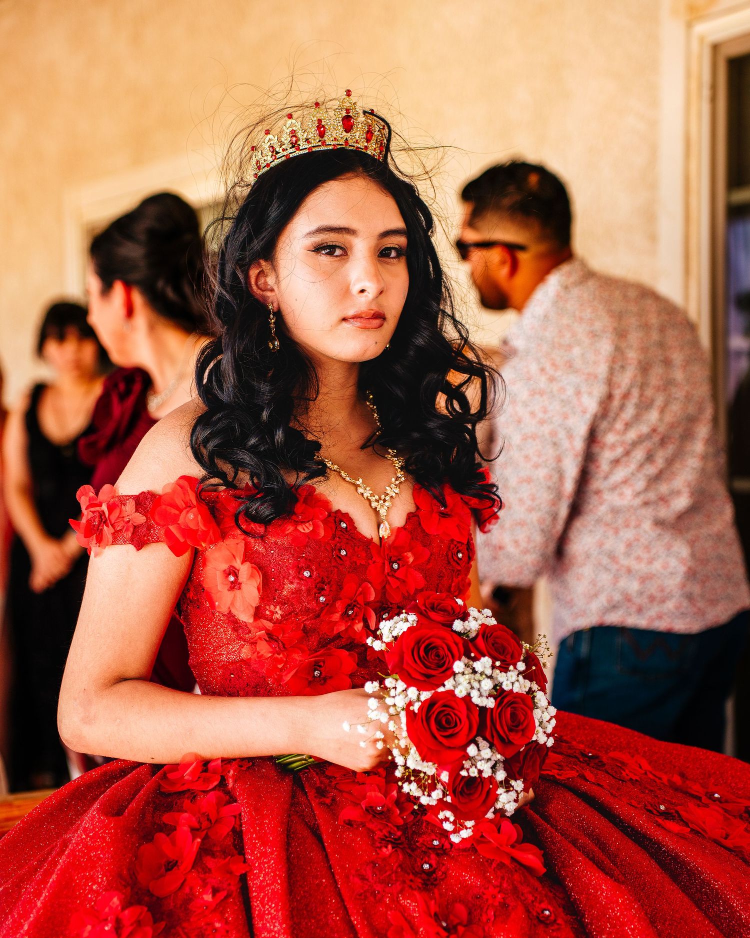 A person in a flowing red ballgown with ruffles poses with a bouquet of red roses against an ornate interior backdrop.