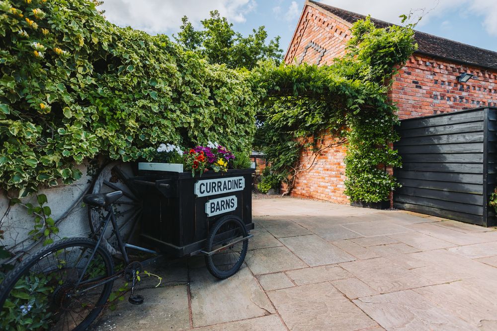 Panoramic view of a garden path with brick walls, climbing vines, and a black wooden gate in the background.