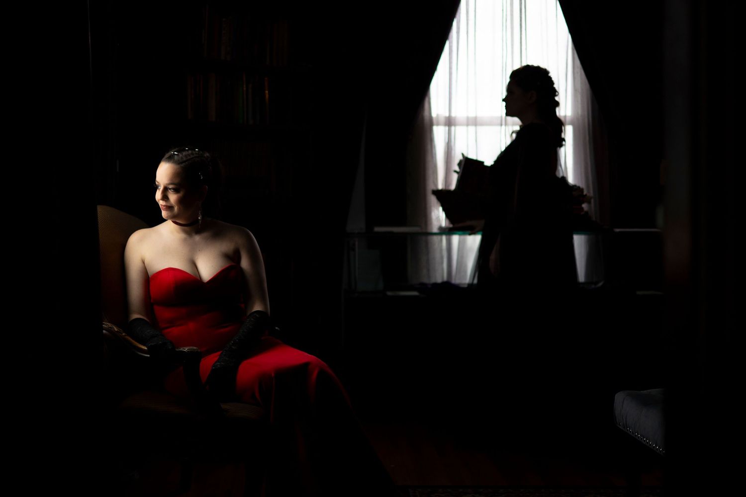 Dramatic noir-style photo of a woman in a red dress seated while a silhouette figure stands by window in the Satanic Temple's library.