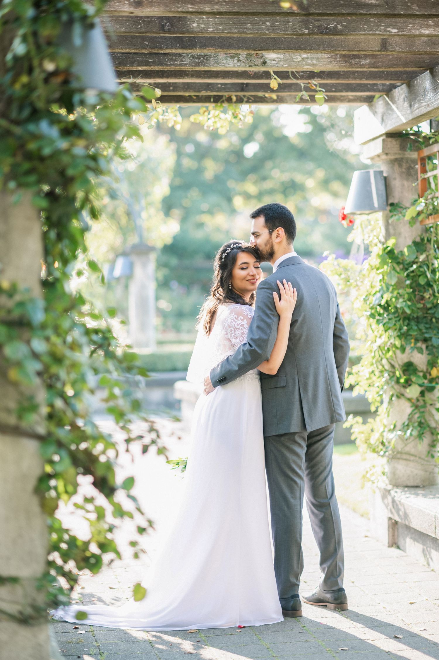 Bride and groom share a romantic embrace under a vine-covered pergola in a lush garden wedding setting.