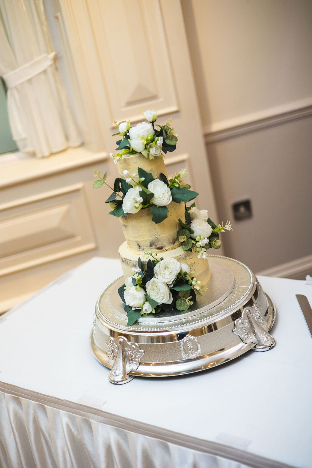 Wedding cake decorated with elegant white roses and greenery displayed on a silver platter.