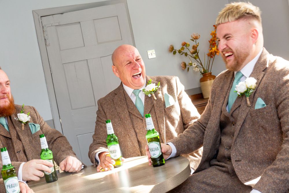 Groomsmen in brown suits share laughs and drinks during a wedding celebration in bright sunlight.
