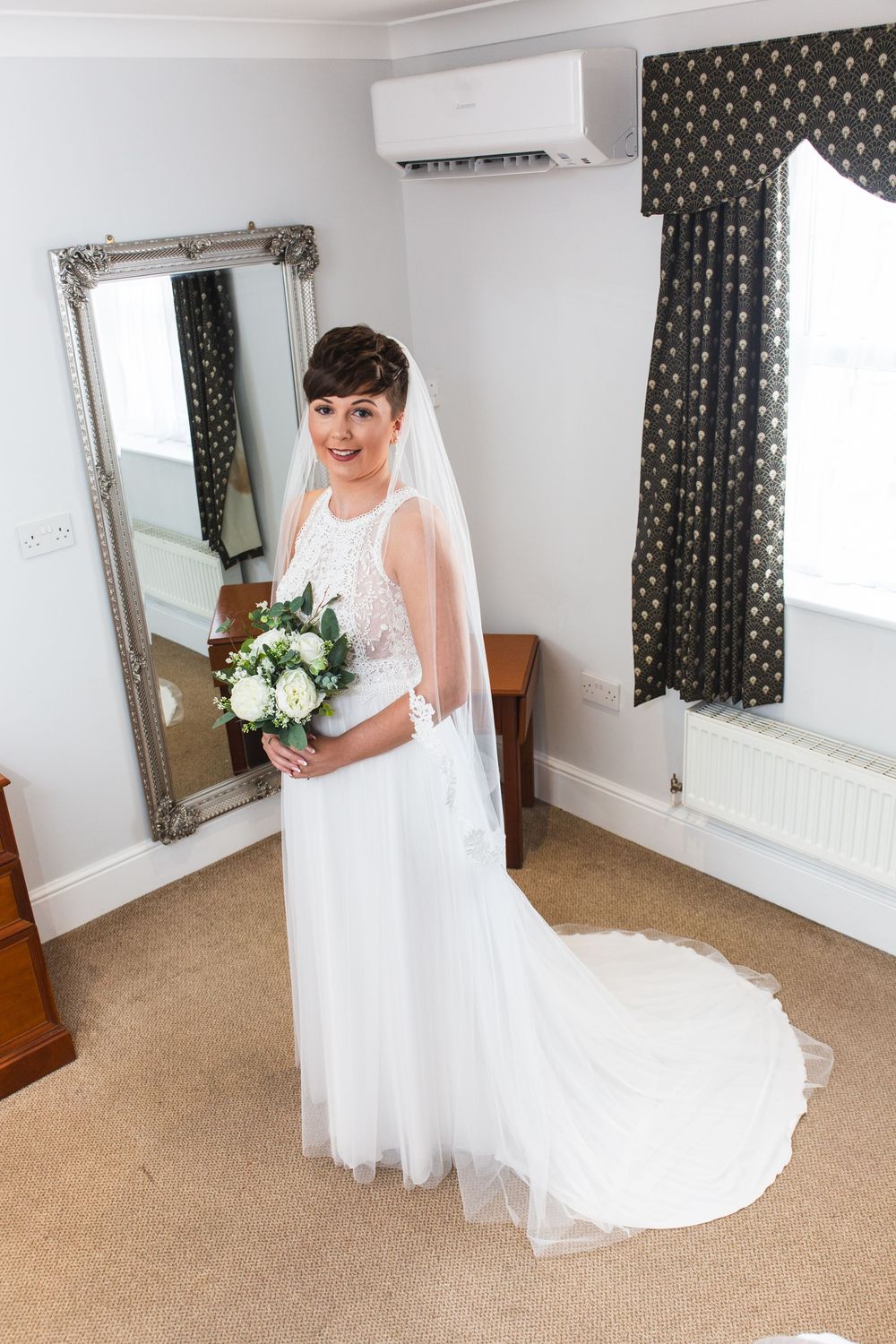 Full-length view of a flowing white wedding dress with tulle skirt in a room with patterned curtains.