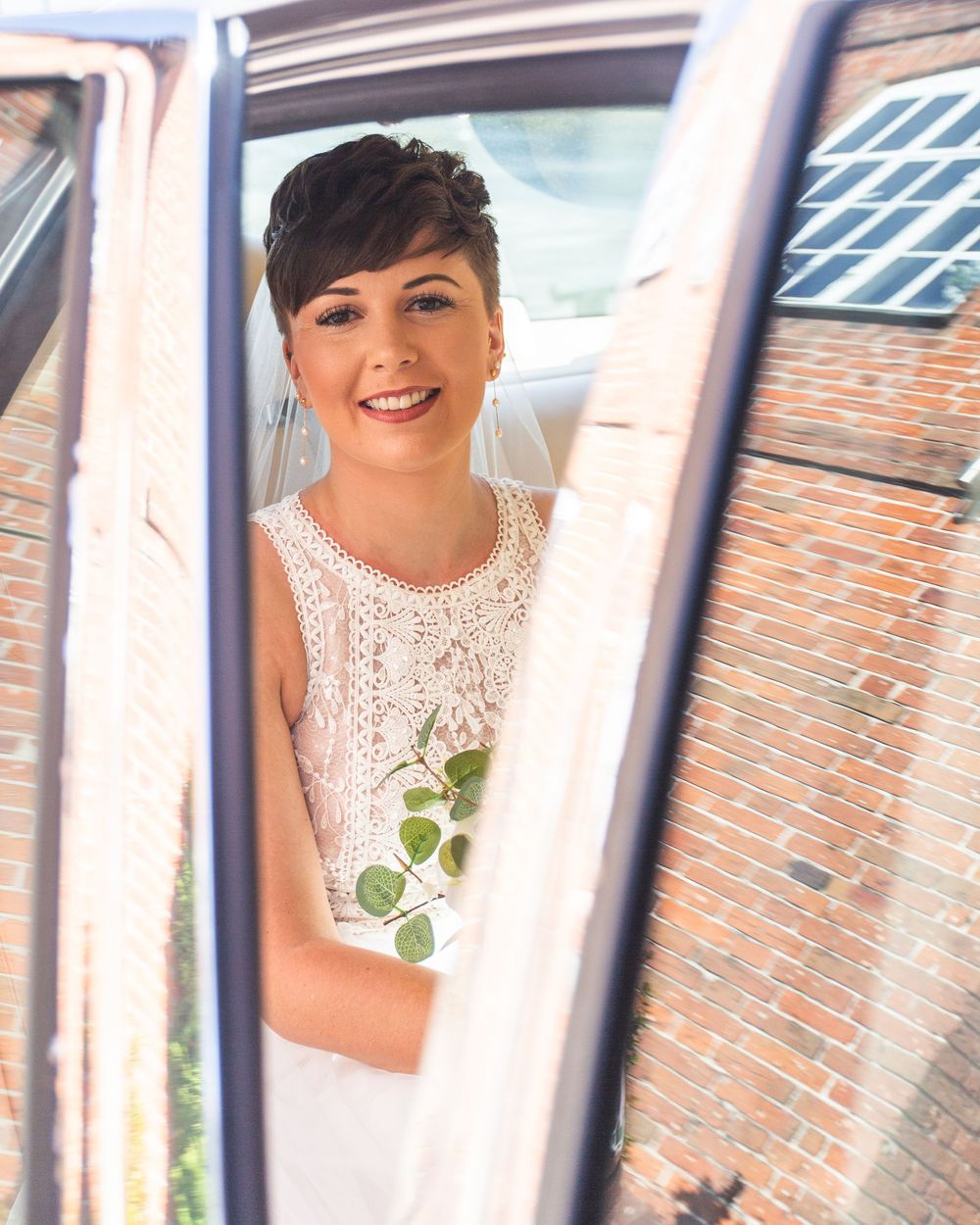 A bride in a white lace dress poses by a window with natural light streaming in, creating an elegant bridal portrait.