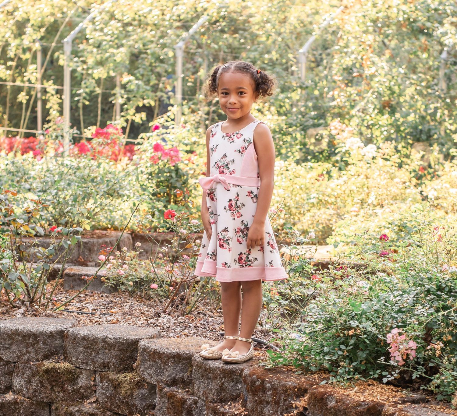 A young child in a floral dress walks along a garden path surrounded by blooming flowers and greenery.