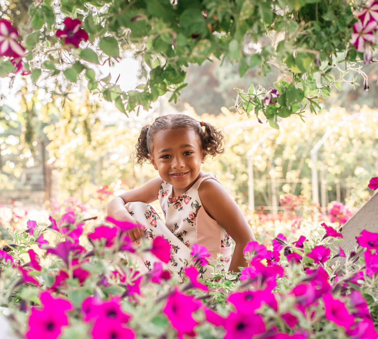 A young child in a white dress poses joyfully among vibrant pink bougainvillea flowers in a sunny garden setting.