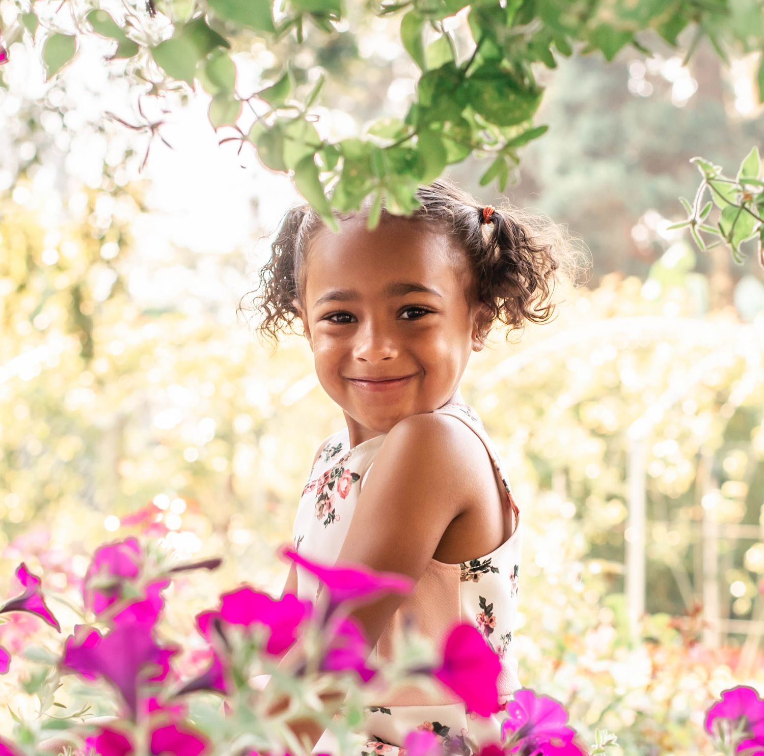 A child peeks through vibrant pink bougainvillea flowers in a sun-dappled garden setting.