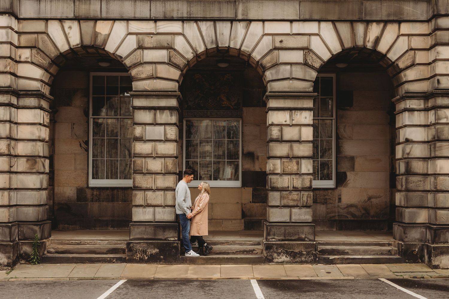 A couple stands together beneath stone arches of a historic building with classic architectural details.