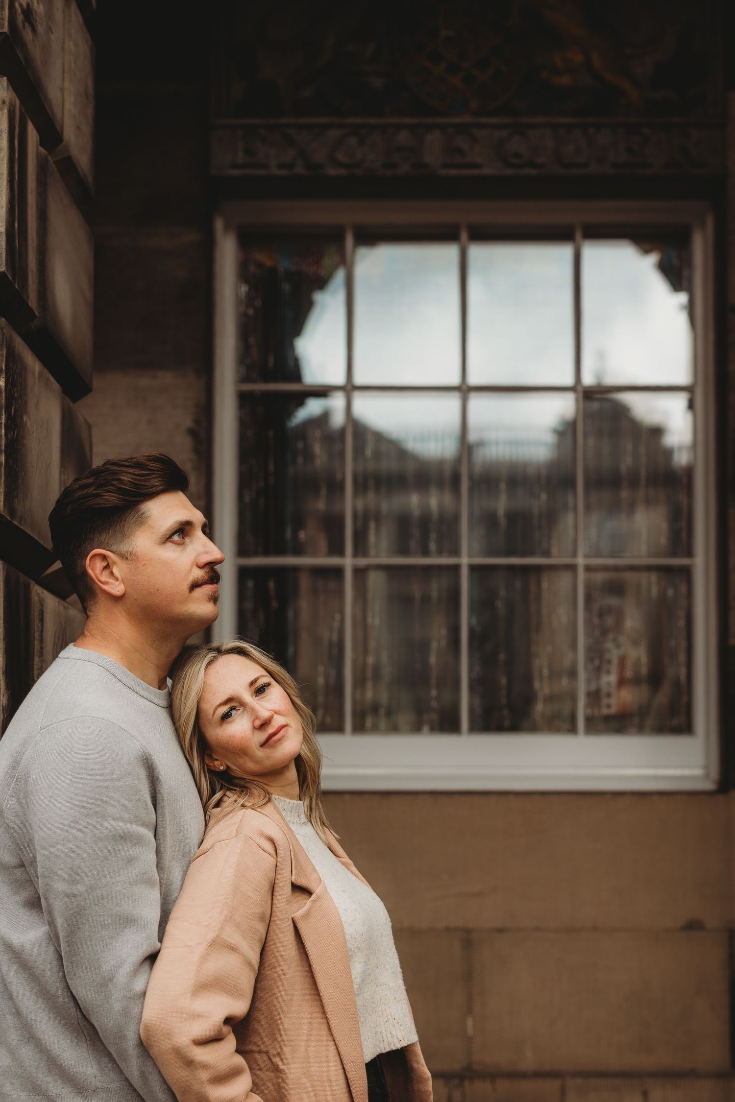 A couple in casual attire poses together in a rustic industrial setting with large windows.