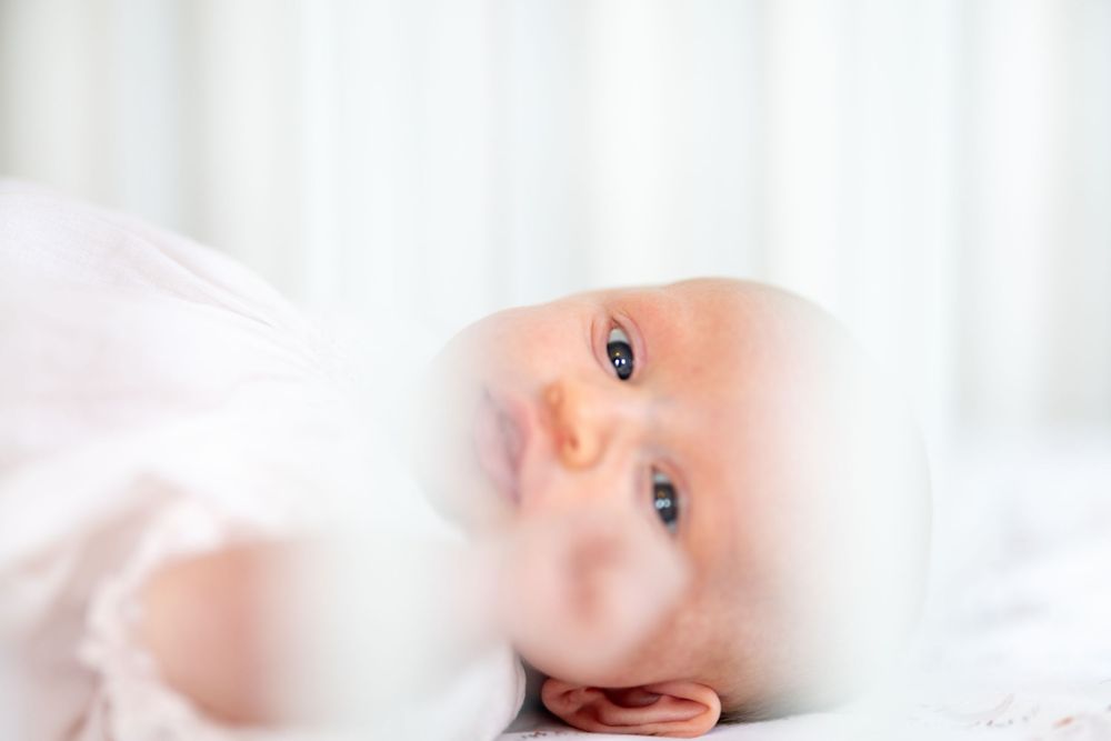 Adorable newborn baby with bright blue eyes gazes upward while lying on white bedding.