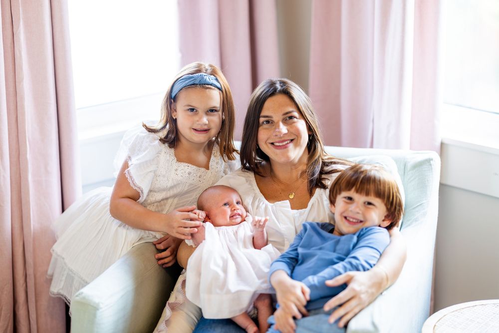 Family sitting together on white couch by window with pink curtains, including newborn baby in white dress.
