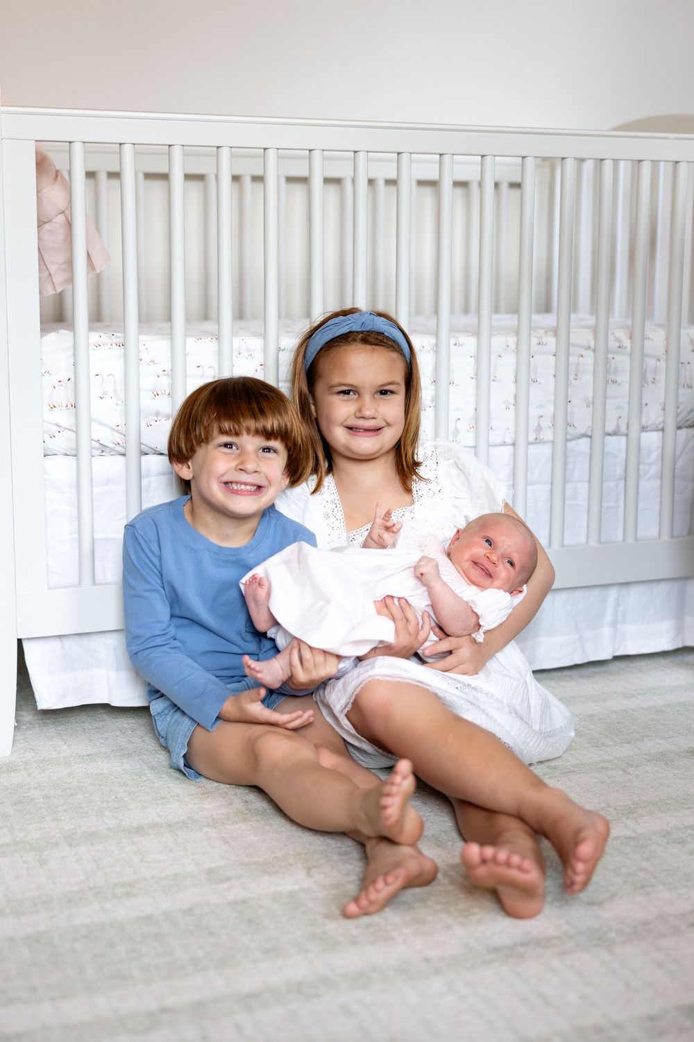 Children sitting on floor with newborn baby in white crib room showing loving sibling bond.