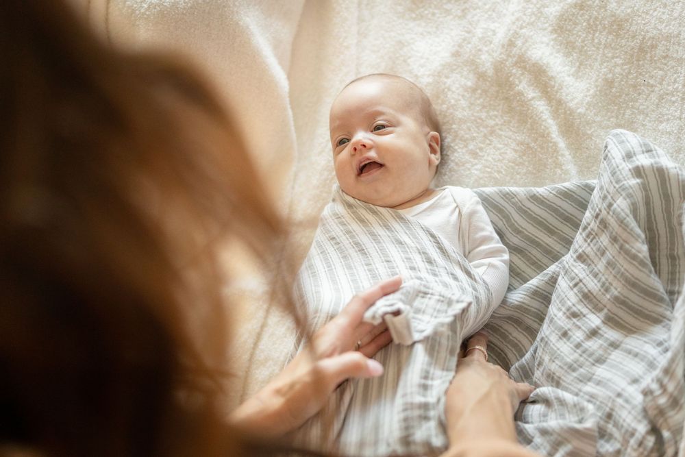 A happy baby wearing a white sweater lies on a soft blanket while being touched by someone's hand.