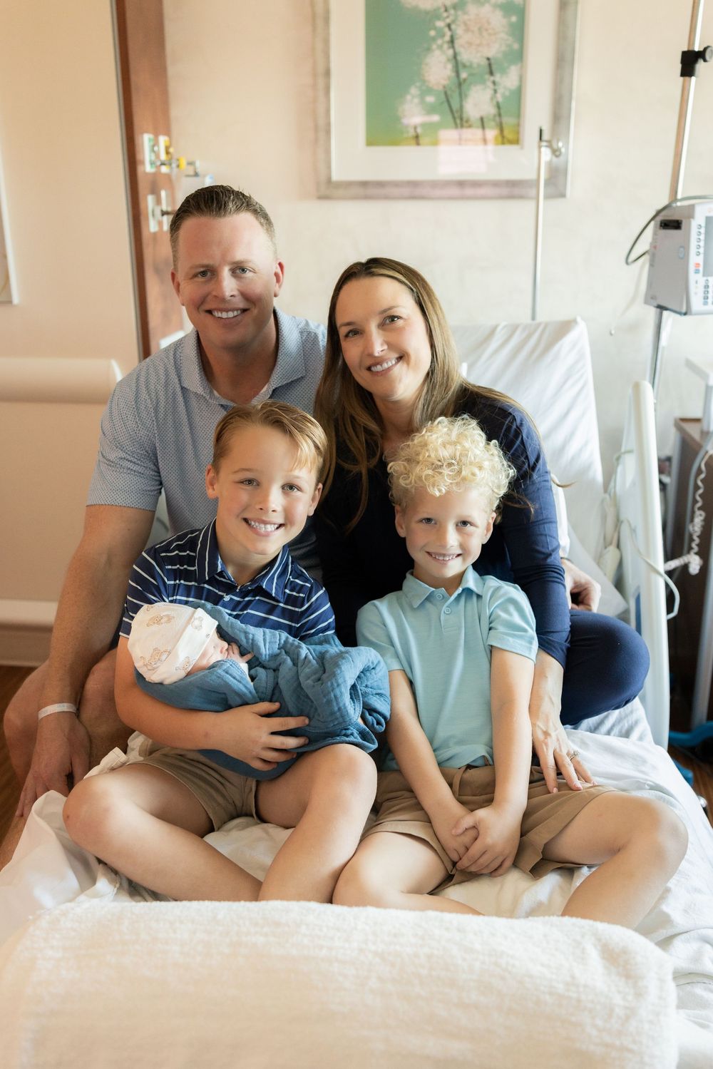 A family sits together on a hospital bed in a medical room with floral artwork on the wall.