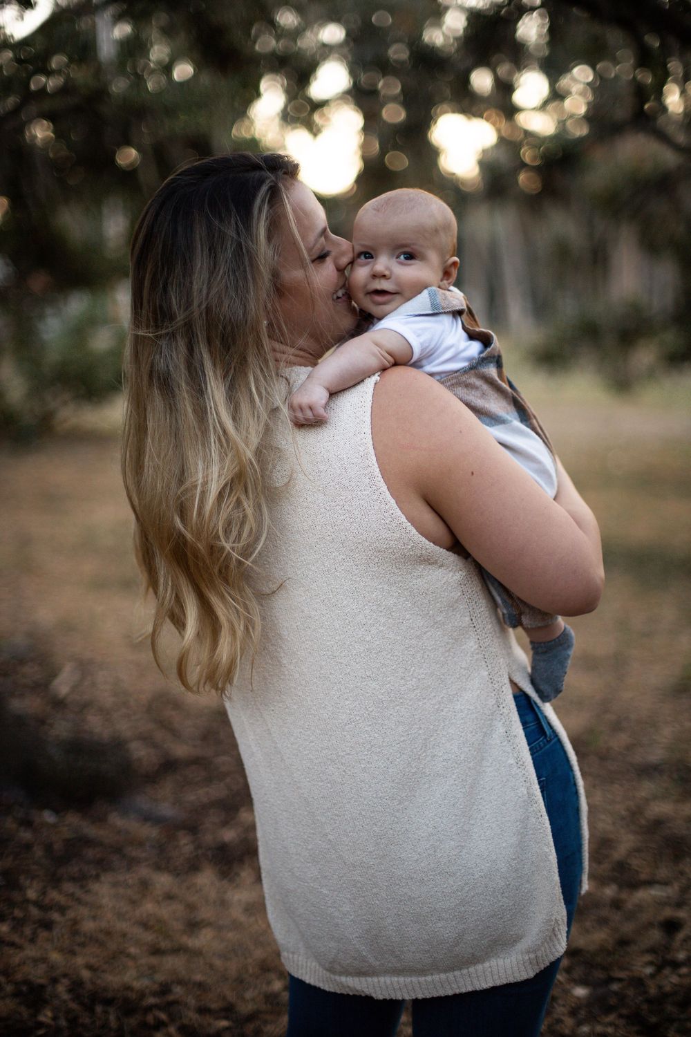 A tender moment captured at sunset as a parent holds and kisses a smiling baby in a white tank top and jeans.