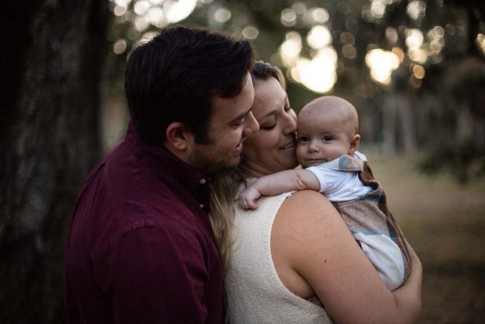 A tender family moment captured outdoors at sunset with parents embracing their baby in a natural setting.