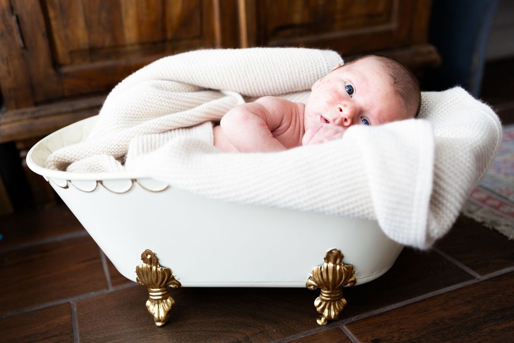 A baby swaddled in white blankets rests peacefully in a decorative white bathtub with gold accents.
