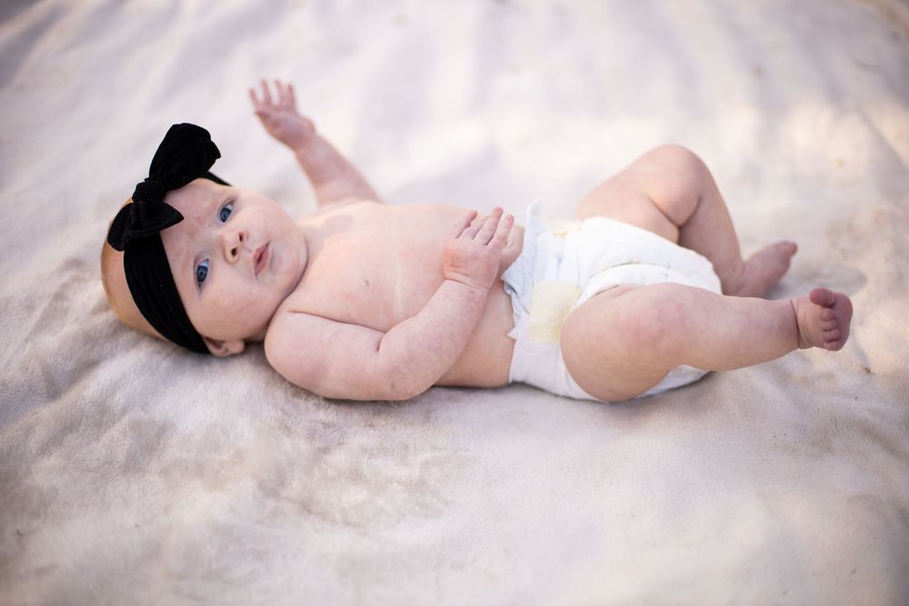 Baby wearing a black headband lies on soft beige fabric wearing a white diaper.
