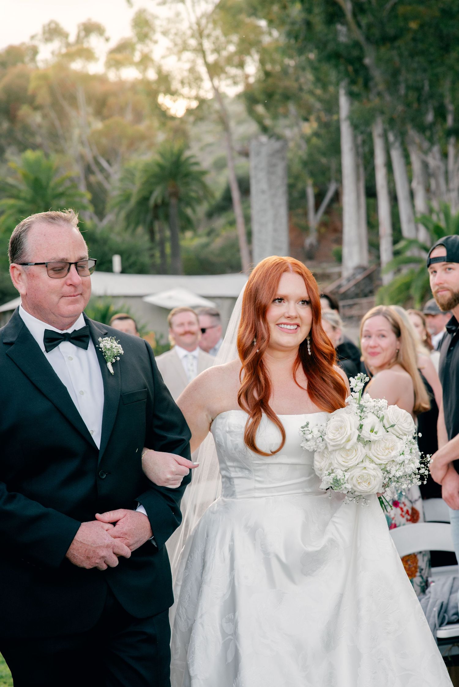 A bride in a white off-shoulder wedding dress walks with her escort at an outdoor wedding ceremony.
