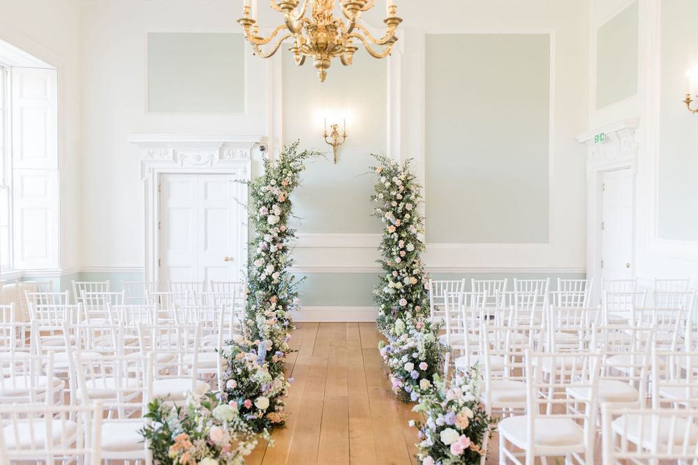 Elegant wedding ceremony setup with white chairs and floral arch in a light-filled room with chandelier.