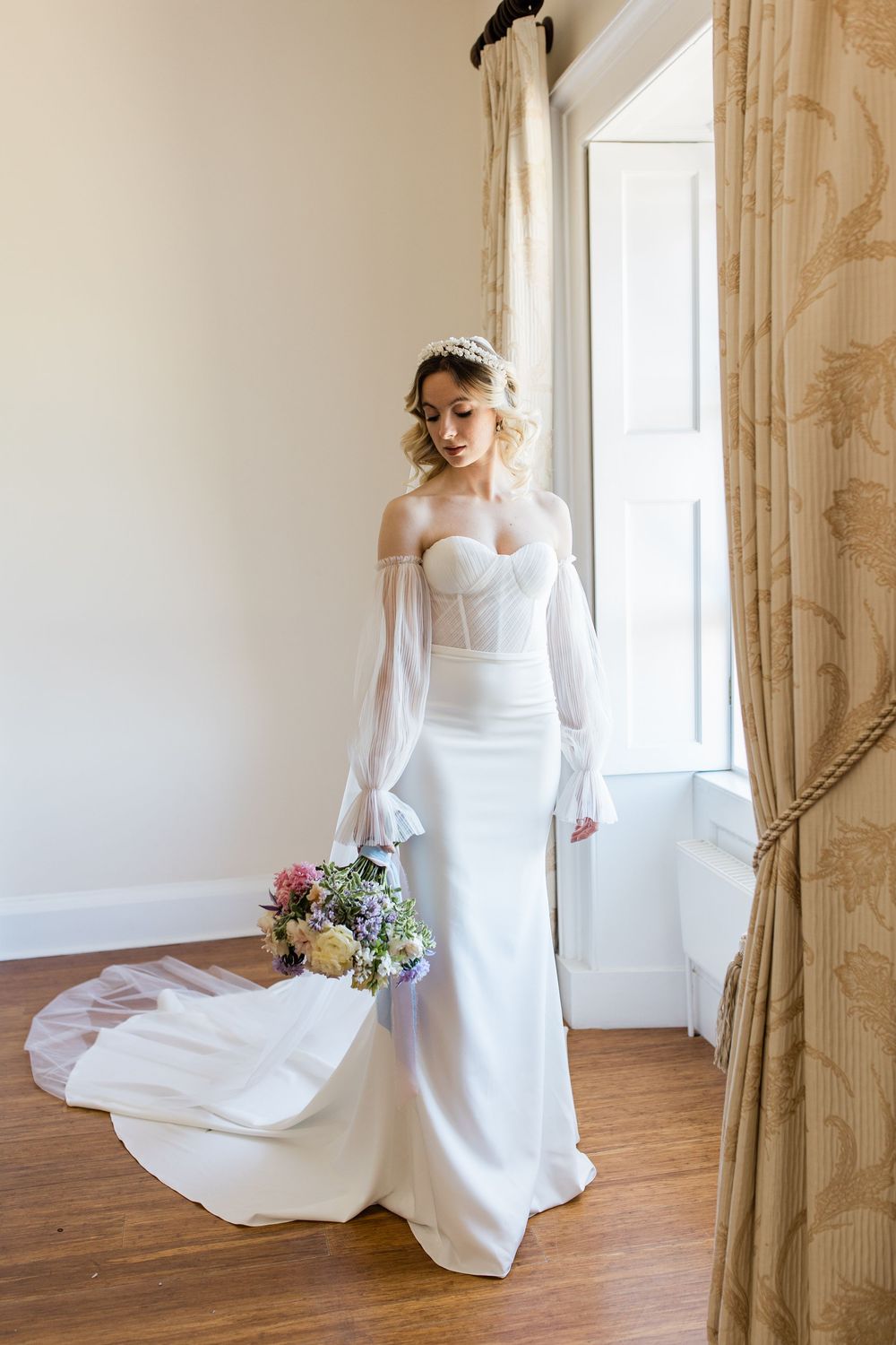 Elegant bride in flowing white dress with long sleeves poses gracefully in a sunlit room with hardwood floors.