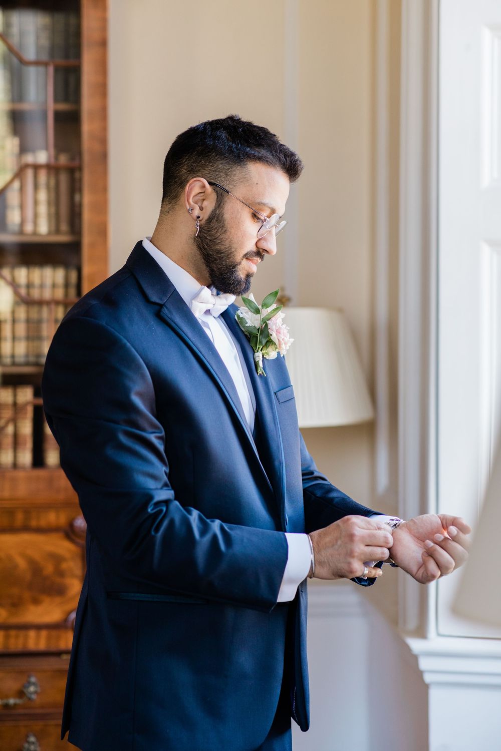 Groom in navy suit adjusts his cufflinks near a bright window.