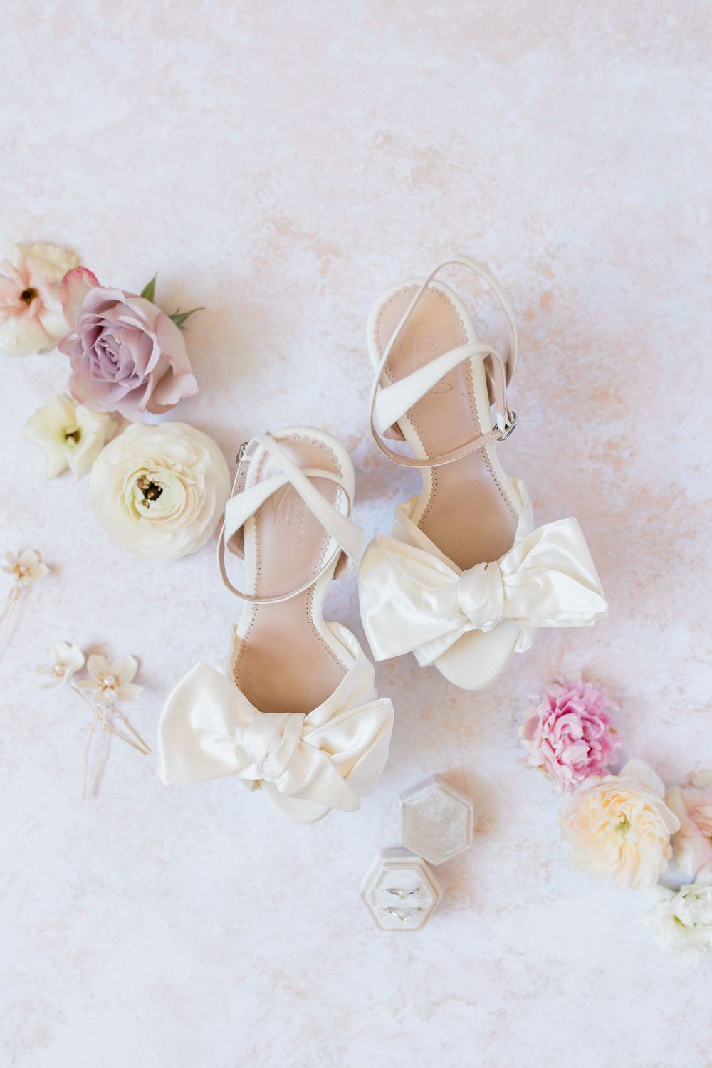 Elegant white bridal shoes with bow details arranged with soft pink and cream flowers on light textured background.