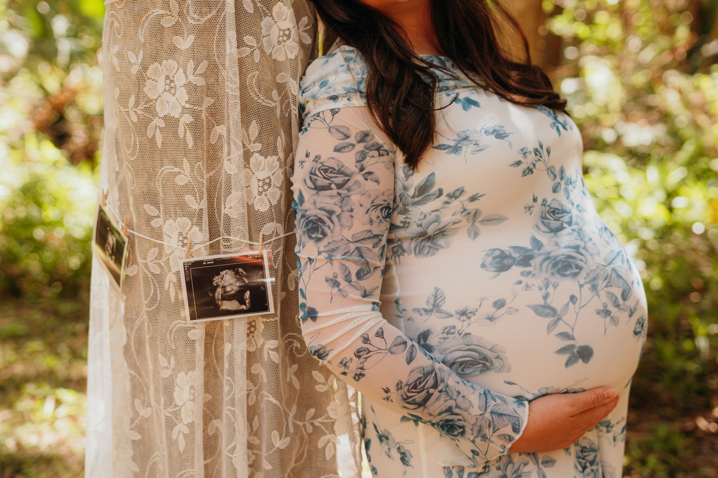 A pregnant woman in a blue and white floral dress poses in a sunlit outdoor setting with trees and foliage.
