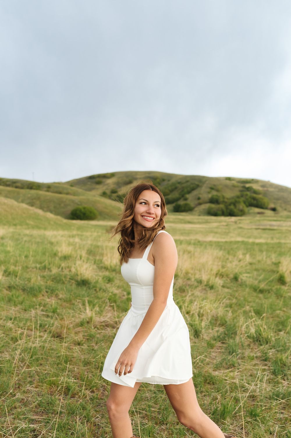 A person in a white dress stands against rolling green hills on an overcast day in a natural setting.