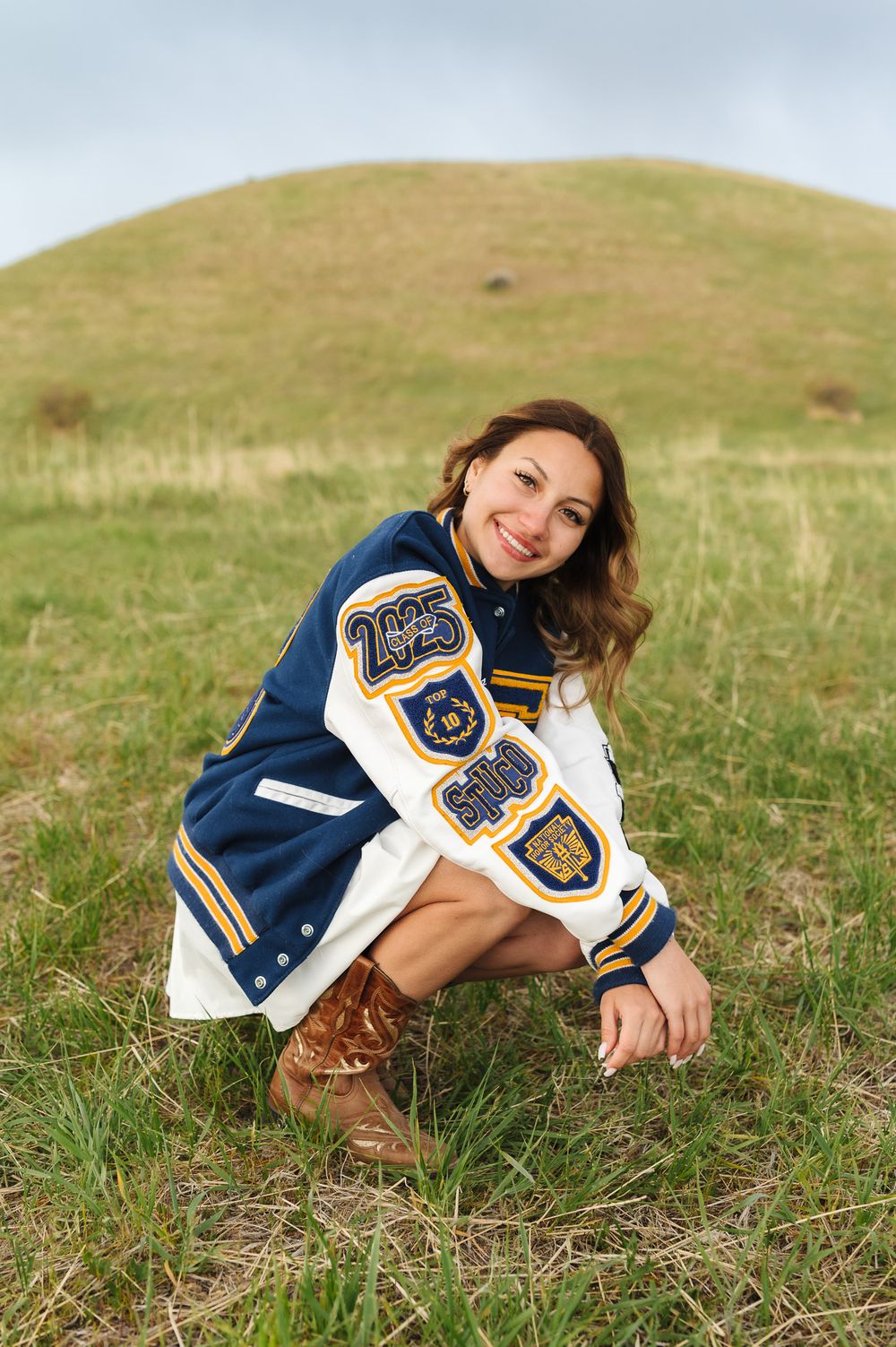 Person wearing varsity letter jacket and blue graduation gown reclines on grassy hillside.