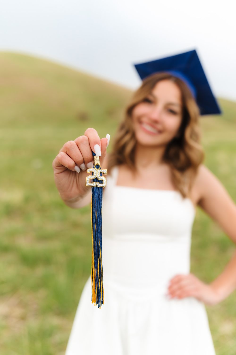 A graduate holds up their tassel and diploma while posing in a white dress against a green outdoor background.