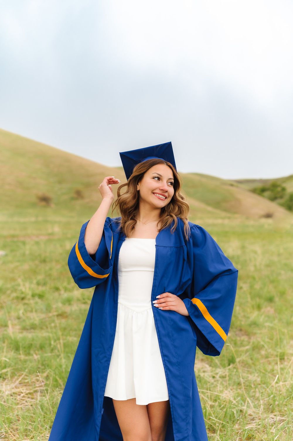 Graduate in blue cap and gown poses playfully in a grassy field against rolling hills at Tunnel Springs