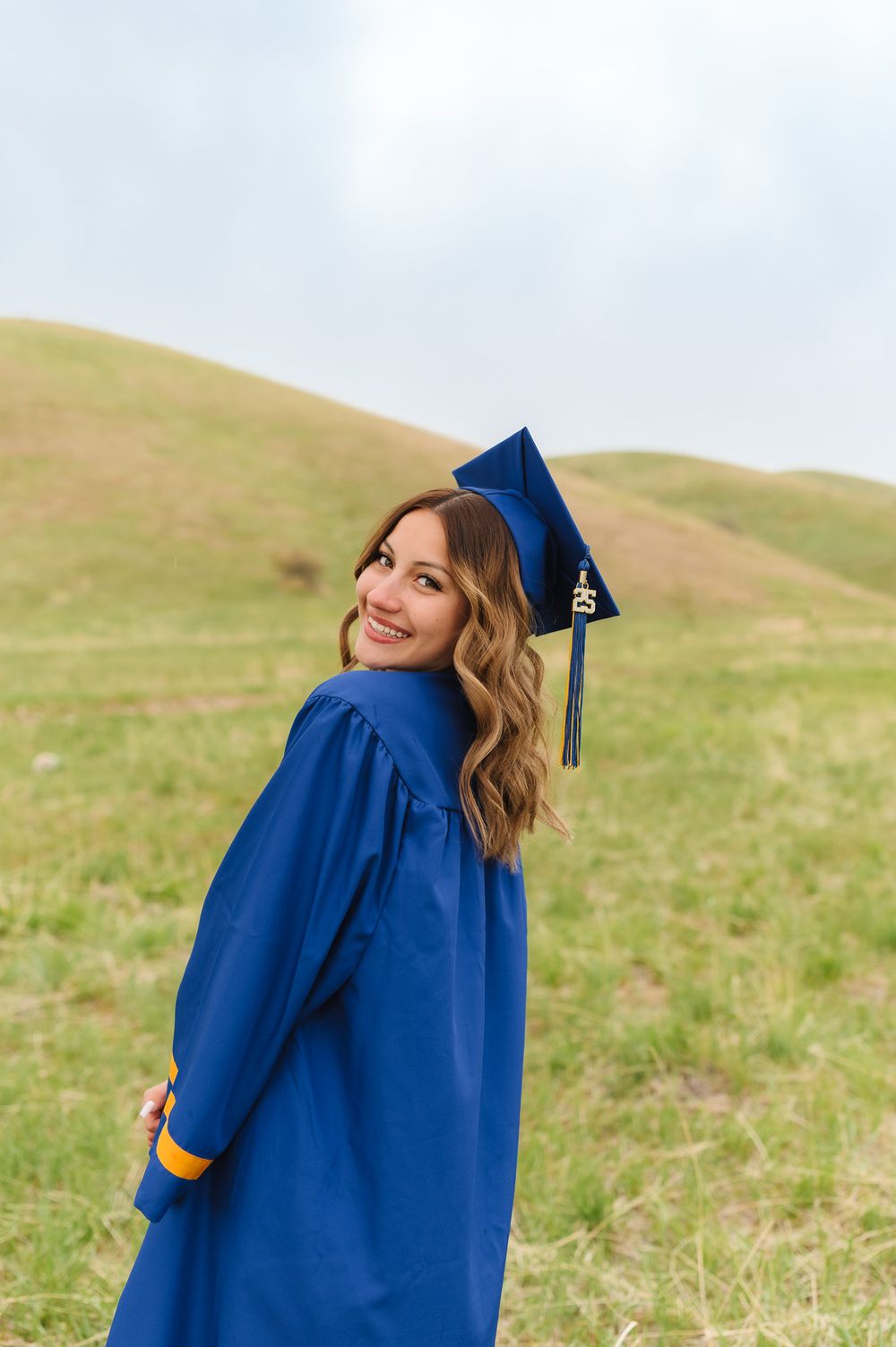 Graduate in blue cap and gown smiles while posing against rolling green hills on a cloudy day in North Salt Lake City, Utah