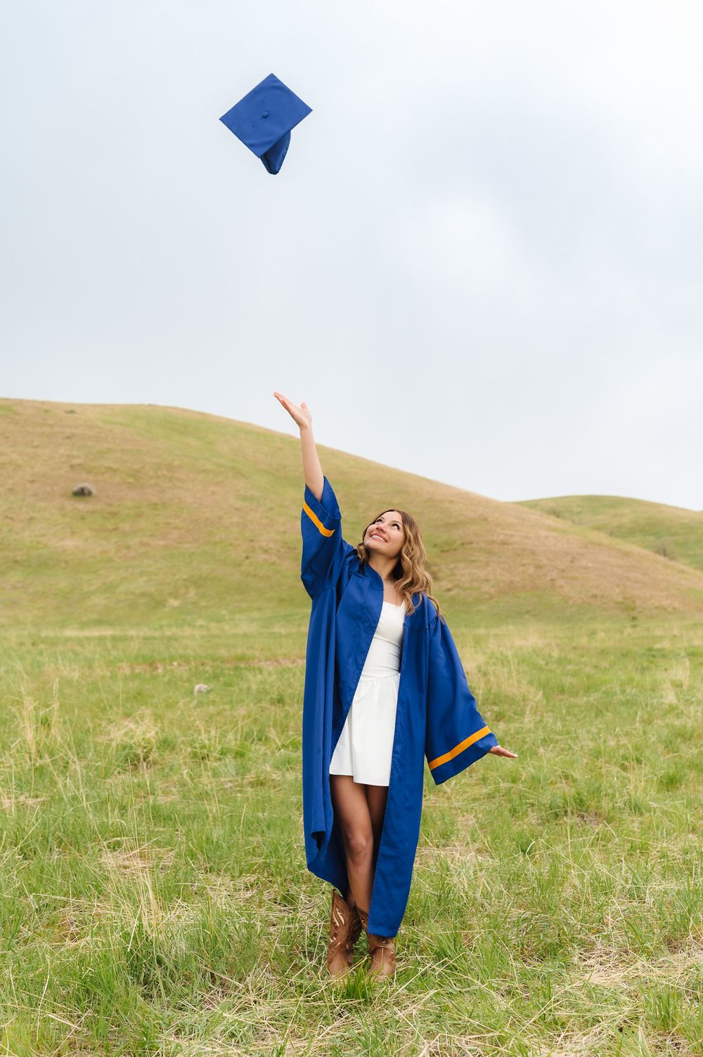 Graduate tossing blue cap in the air while celebrating on a hillside under cloudy skies at Tunnel Springs, Utah.
