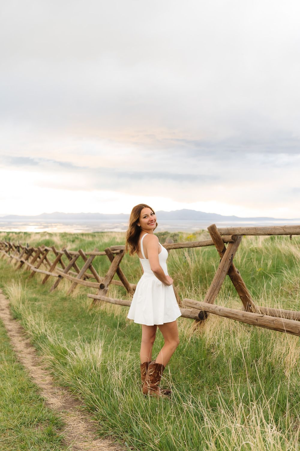 A white sundress and brown cowboy boots complement the rustic wooden fence and mountain landscape at sunset at Tunnel Springs, Utah