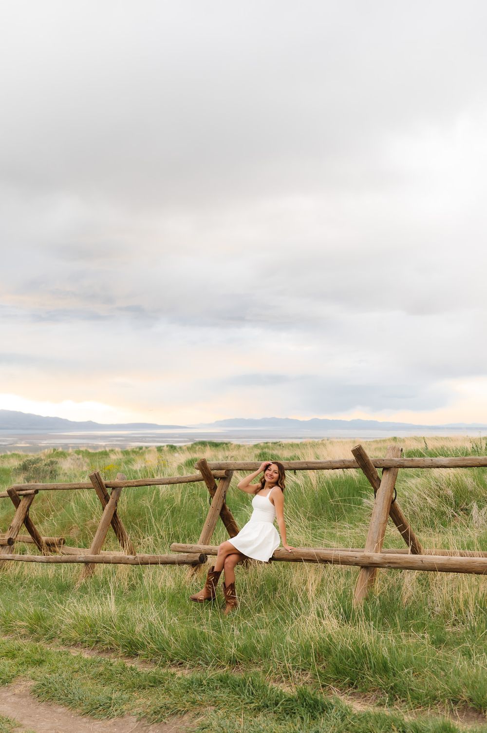 Person lounging on rustic wooden fence in countryside with mountains and cloudy sky in background.