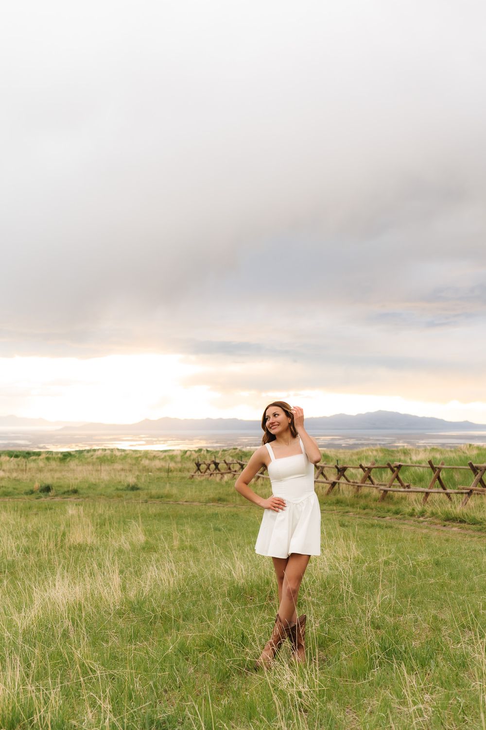 Graduate in white dress poses joyfully in a grassy field with mountains and sunset in the background.
