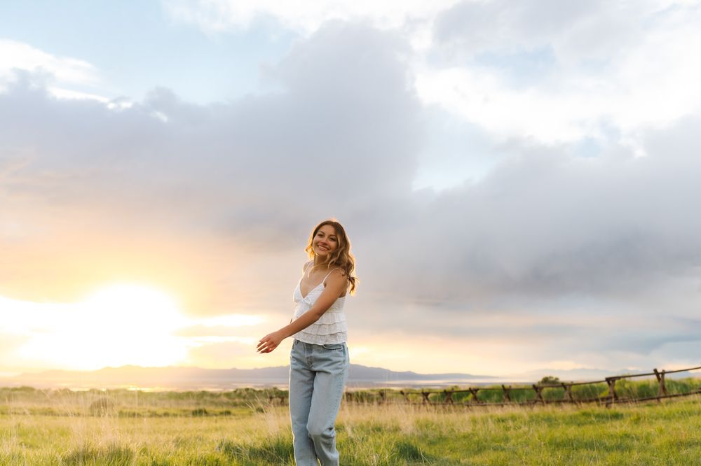 A person spins joyfully in a grassy field at sunset with dramatic clouds and golden light in the background.