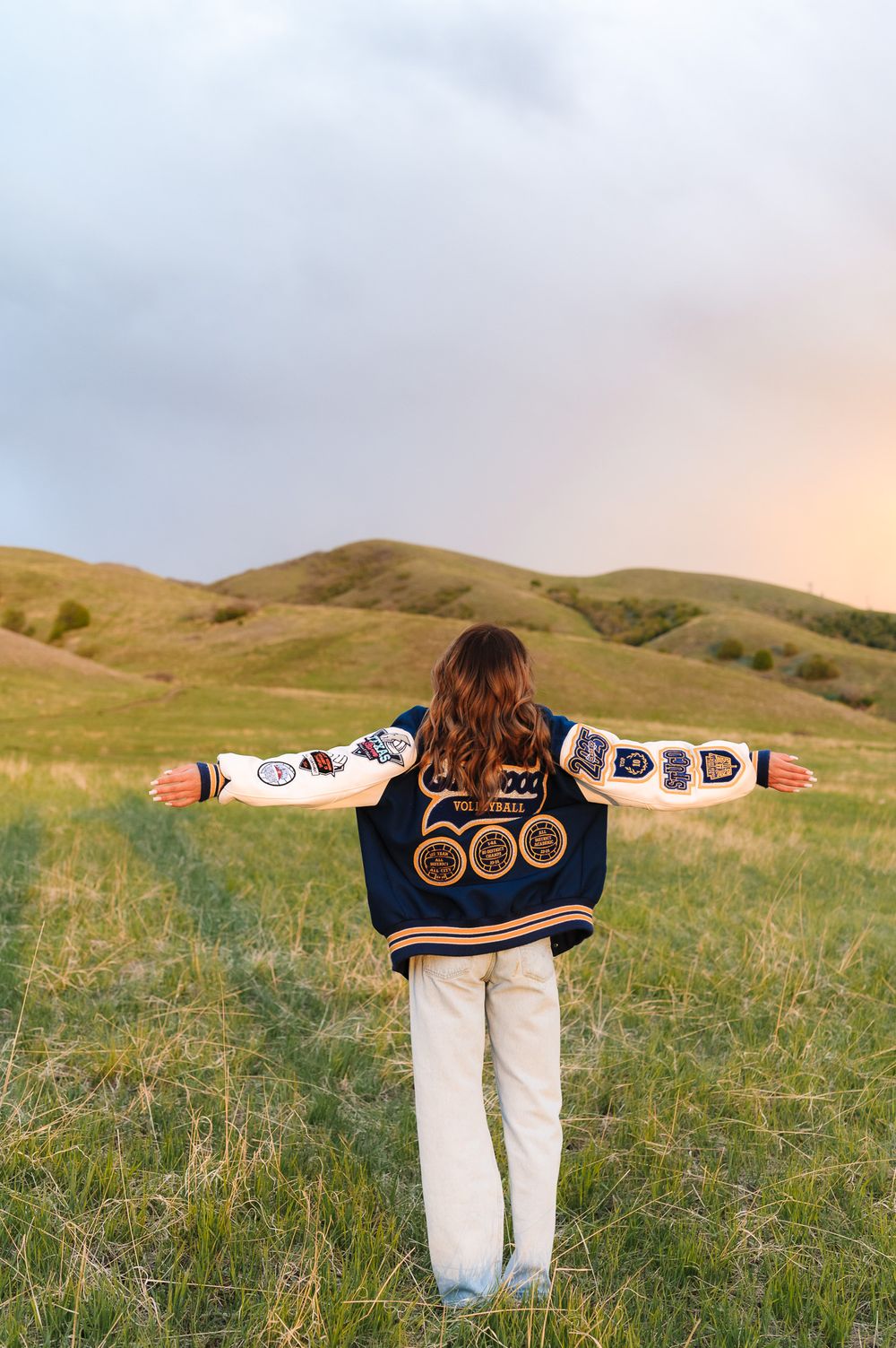 Person wearing embroidered jacket with arms outstretched stands in rolling green hills at sunset.