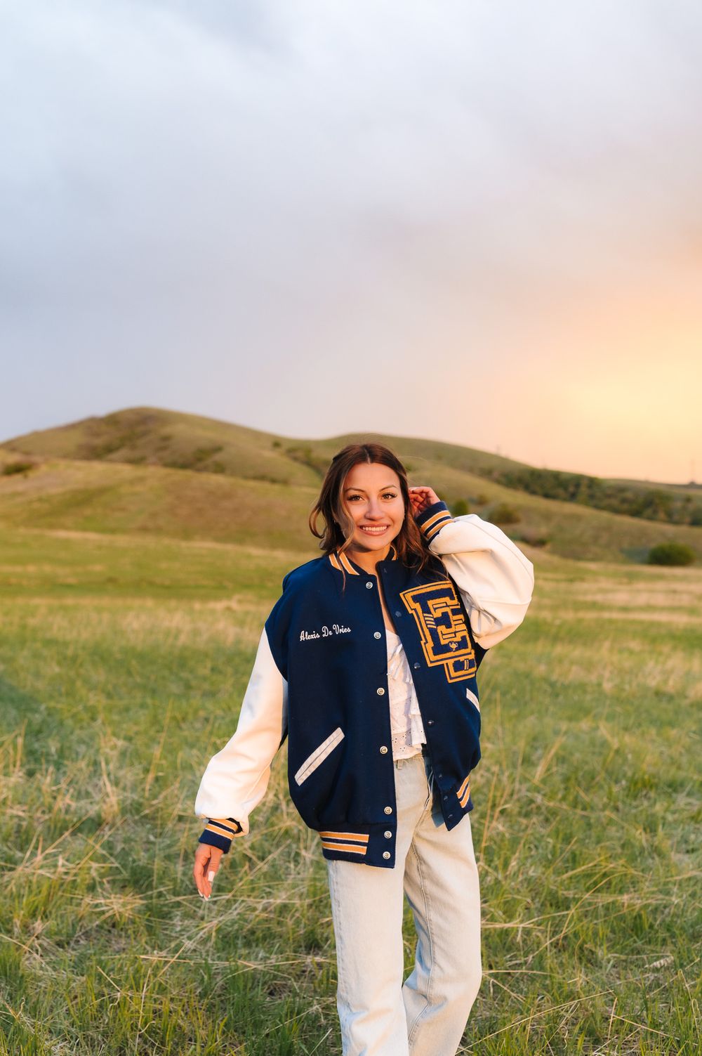 Senior session of a girl wearing aVarsity letterman jacket and white pants outfit styled in golden hour lighting against rolling green hills