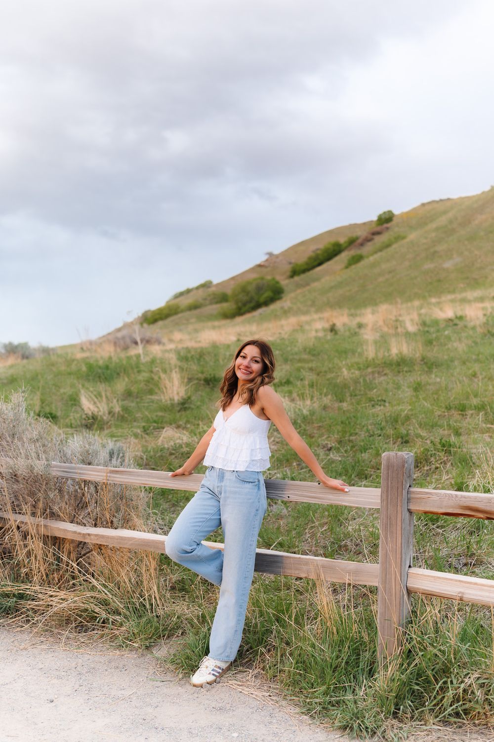 Person in light top and jeans poses playfully along wooden fence on grassy hillside at sunset.
