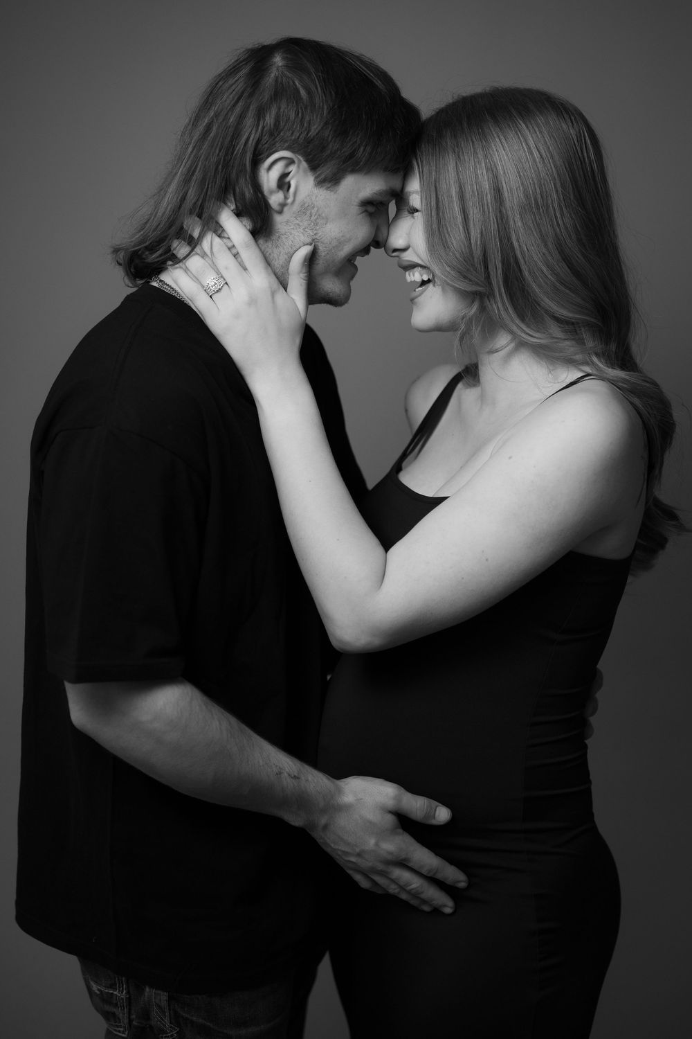 Black and white portrait of an intimate couple embracing, with the woman wearing a tank top and showing a pregnant belly.