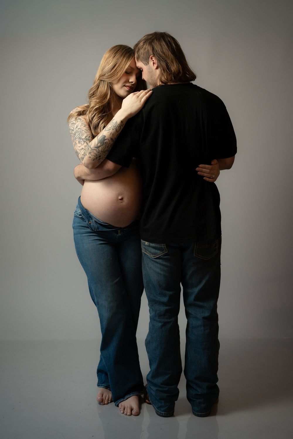 Intimate maternity portrait showing a couple embracing in casual attire against a neutral backdrop.