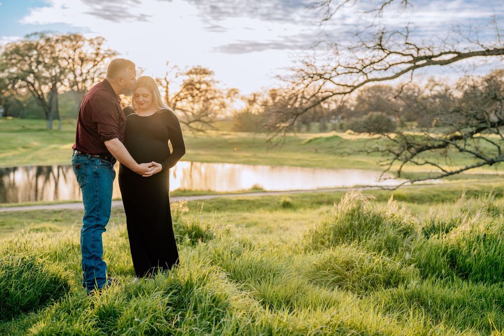 A couple stands together on a grassy hill at sunset with a tranquil pond in the background.
