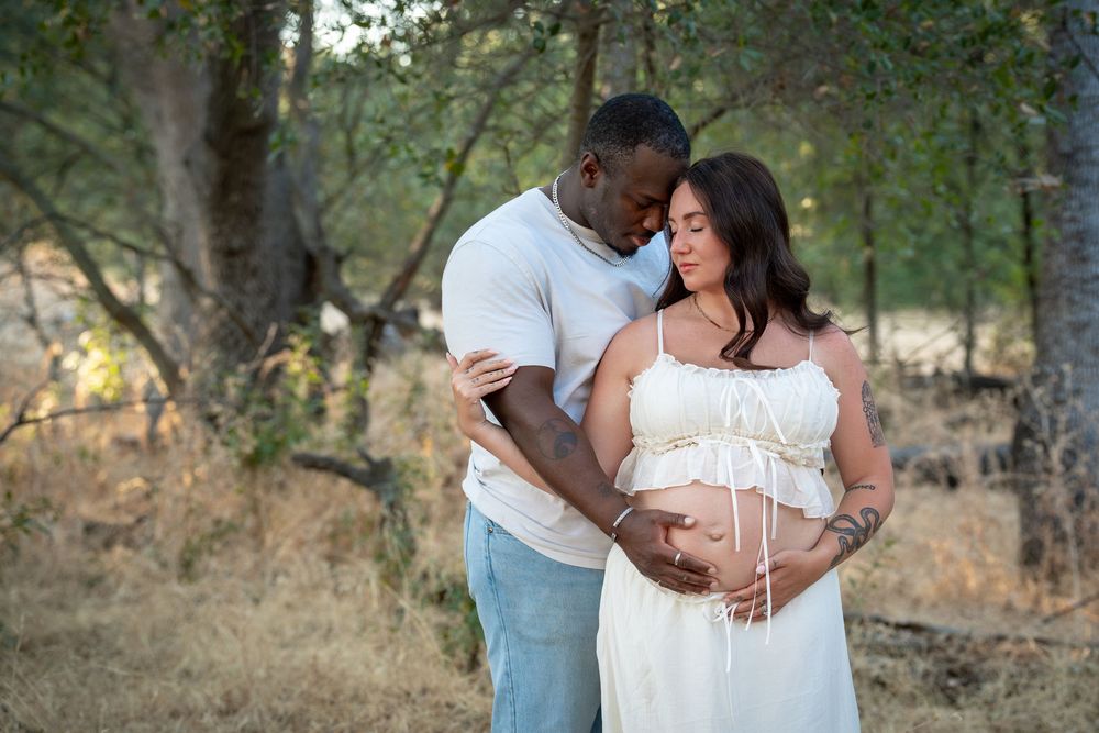 Couple sharing a tender maternity photo outdoors, with man embracing pregnant woman in white dress among trees.