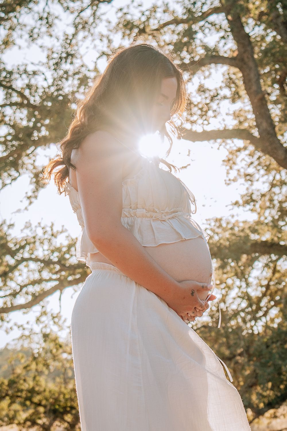 Pregnant woman in white dress cradling baby bump outdoors with sun flare through trees.